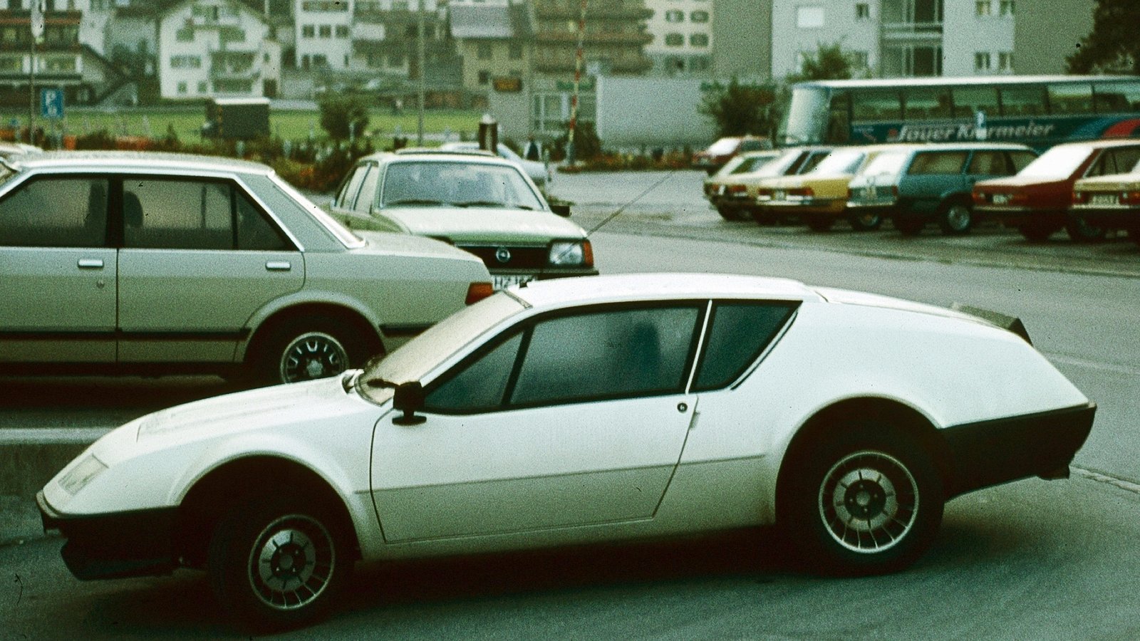 White early-1980s Alpine A310 V6 coupe in side profile, showing its wedge nose, glassy rear hatch and wide rear wheel arches, parked on a street in the Alps