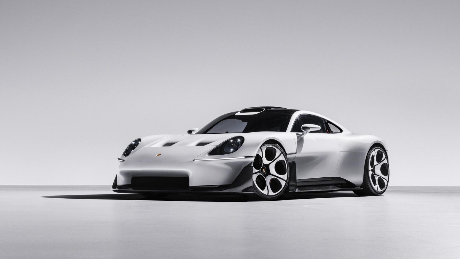 Front three-quarter view of the white Porsche Mission S in a grey studio, showing round headlights, a low flat nose with dark intakes, a dark glass canopy and large white-and-black multi-spoke wheels