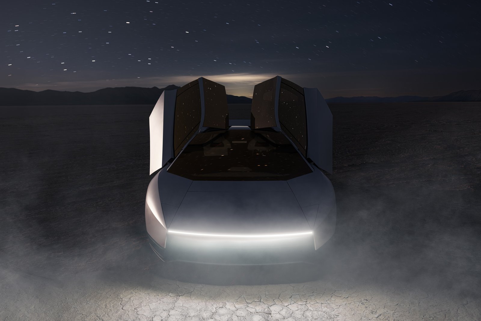 Front view of the Tesla Roadster at night on a cracked dry lakebed under a starry sky, both upward-opening doors raised and a thin full-width light strip glowing across the nose