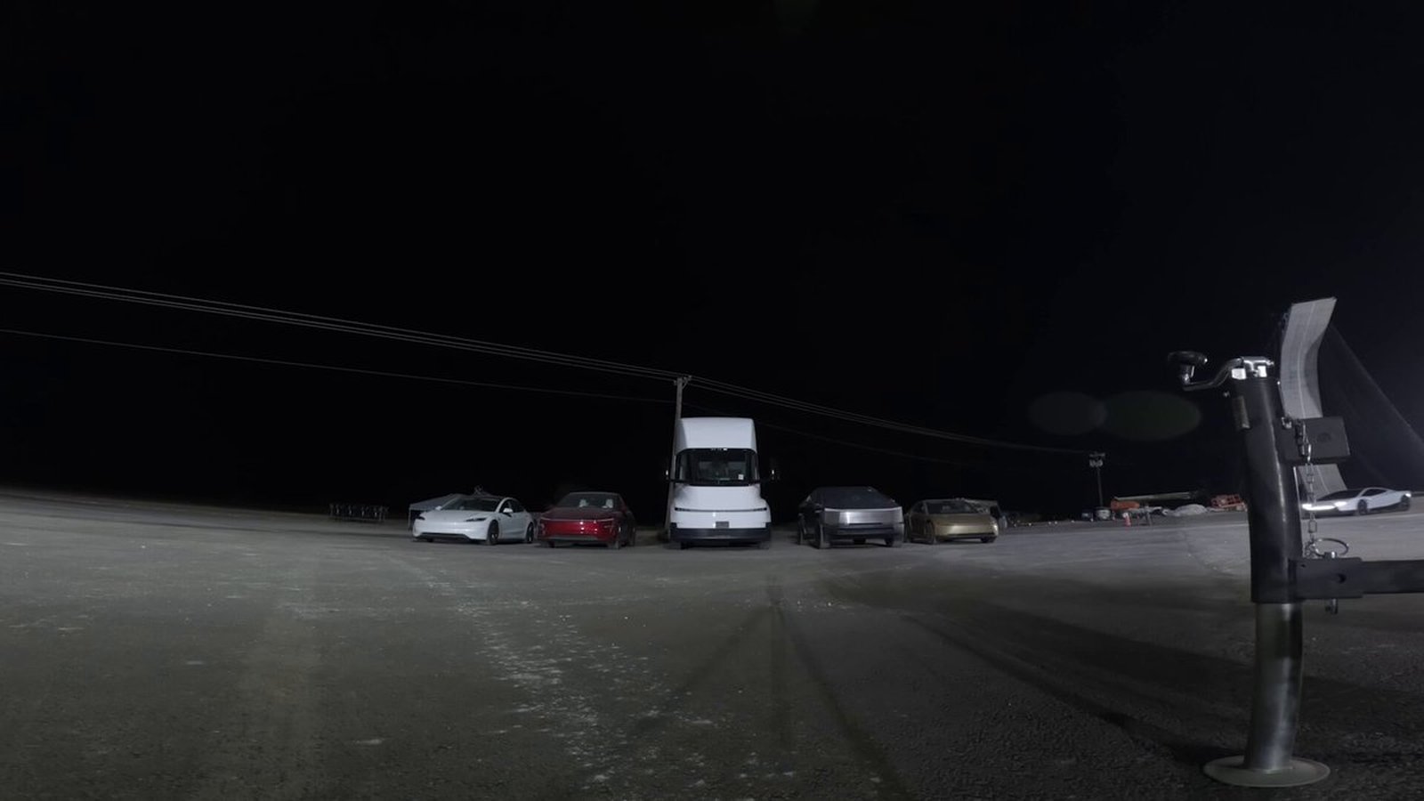 Opening frame of Elon Musk's clip: a white Tesla, a red Tesla, a Tesla Semi, a Cybertruck and a gold Cybercab lined up on a dark dirt lot at night, with a camera rig in the foreground and the white Roadster in the distance at right