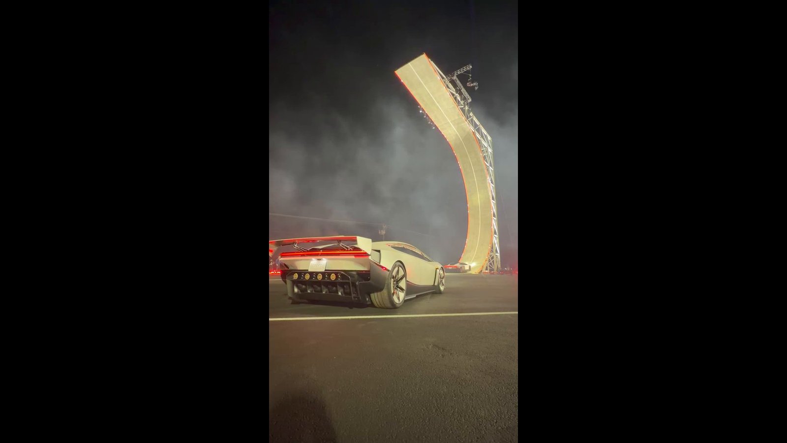 Opening frame of Franz von Holzhausen's vertical video: the white Tesla Roadster seen from the rear with its wing raised and red taillight lit, in front of a tall curved stunt ramp at night