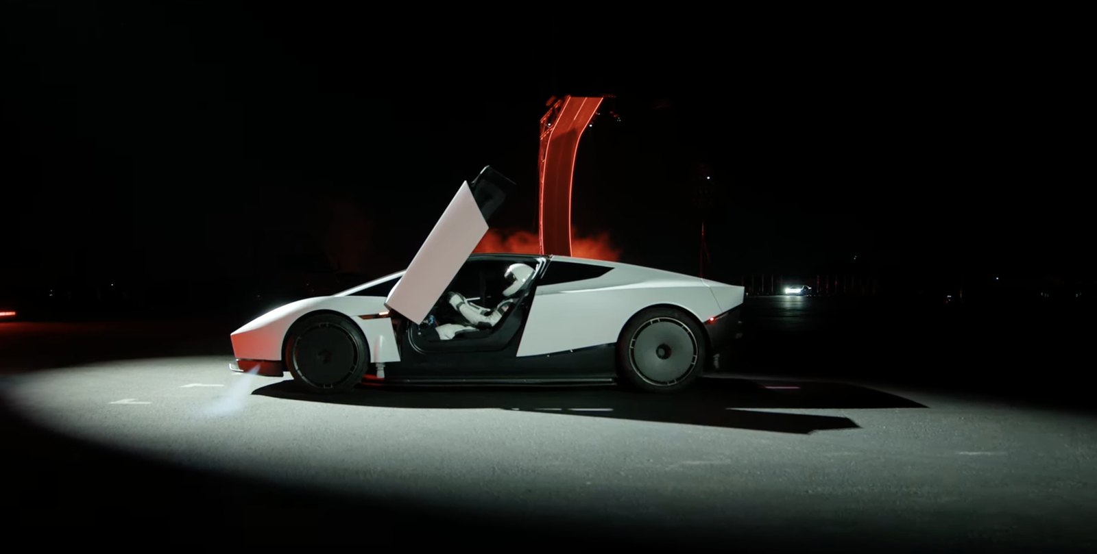 The new white Tesla Roadster in side profile at night under a spotlight, its upward-opening door raised, with a figure in a white spacesuit in the driver's seat and a red structure in the background